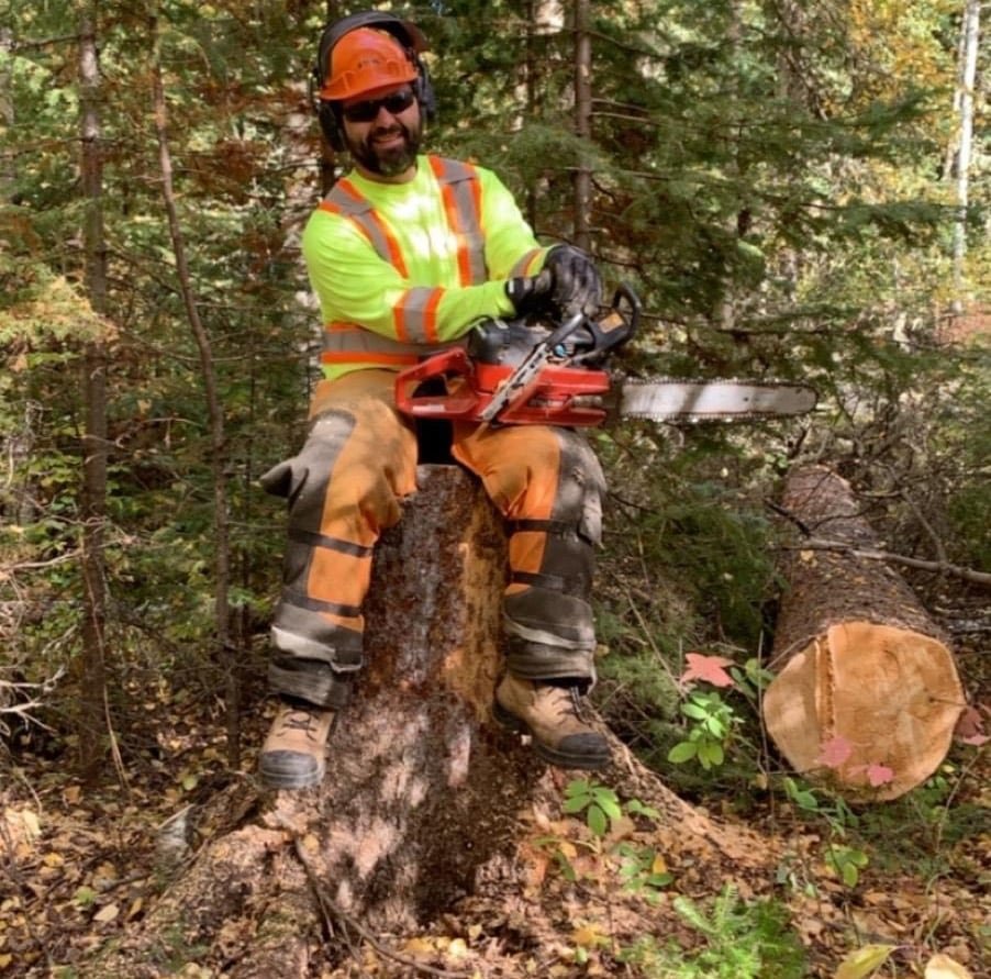 A bearded man in protective gear holding a chainsaw on a tree stump with a fallen tree nearby.