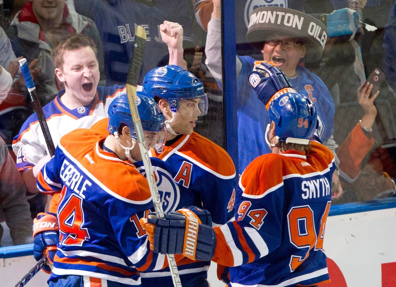 Three hockey players in orange-and-blue jerseys embrace in celebration while fans cheer on the other side of the arena glass.
