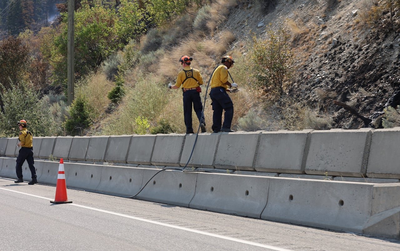 Firefighters in yellow gear working near a highway with charred ground using hoses.